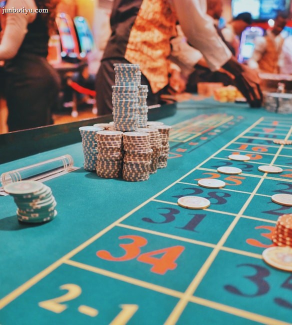 A casino scene with a roulette table features prominently, covered in green felt with colorful numbers and betting squares. Several stacks of poker chips are placed on different numbers, and the hands of several people are visible as they interact with the table. In the background, there are slot machines and more people, creating a vibrant and busy atmosphere.