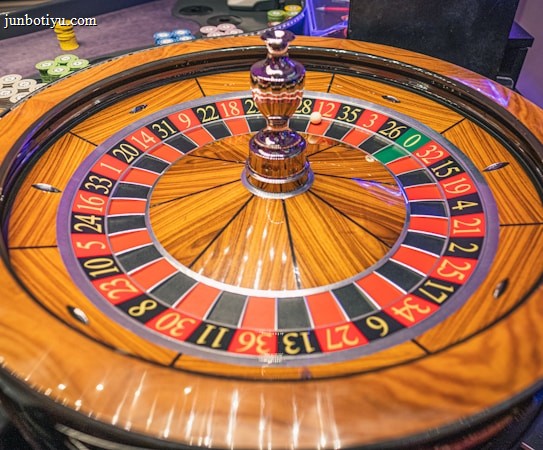 A roulette wheel with alternating red and black numbered pockets on a wooden platform. The wheel contains a small white ball resting in one of the pockets, indicating gameplay. Surrounding the wheel are stacks of colorful poker chips, suggesting a casino environment.