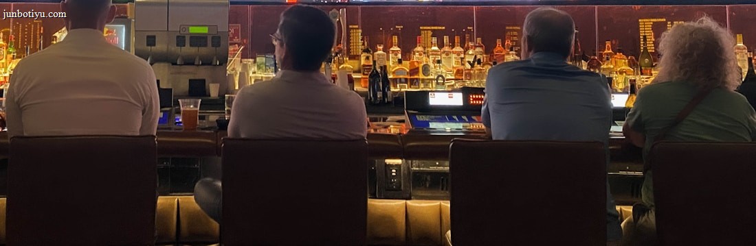 A group of people sit at a bar with their backs to the camera, watching multiple sports games displayed on large screens. The bar counter is lined with various bottles of liquor, and the atmosphere suggests a casual and relaxed social setting.