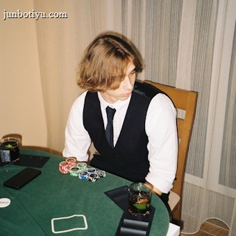 A person with shoulder-length hair is sitting at a poker table. They are wearing a white shirt, black vest, and tie, with an array of poker chips in front of them. There are two glasses with drinks on the table, and a smartphone is placed nearby. The background consists of vertical striped curtains.