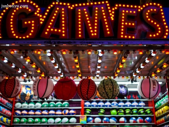 A brightly lit carnival game stall with a large 'GAMES' sign at the top surrounded by red and yellow lights. Various sports balls, including basketballs and soccer balls, are hanging from the ceiling and neatly arranged on shelves below.