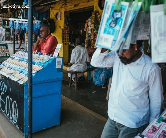 A street vendor stall displaying an array of lottery tickets clipped to a bright blue kiosk. A man in a red shirt leans on the counter, appearing thoughtful, while another man in a white shirt adjusts his hat and walks by. A variety of products hangs from strings above, and a colorful display fills the background with vibrant signs and packets. The scene suggests a busy, lively marketplace.
